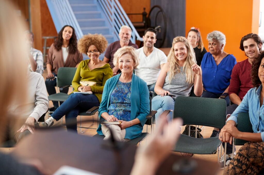 A group of people sit in chairs in a meeting room, listening to a speaker.