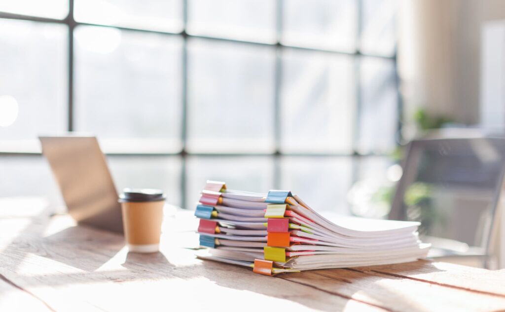 A stack of bound documents on a desk with a laptop and a coffee cup.